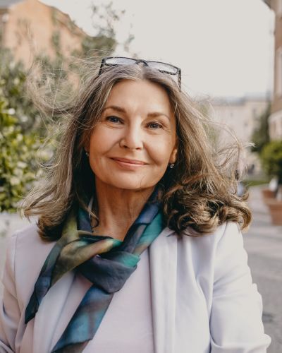 Outdoor image of gorgeous positive lady with charming smile and loose gray hair enjoying nice summer day, wearing white jacket, silk scarf and eyeglasses. Beauty, urban style and fashion concept